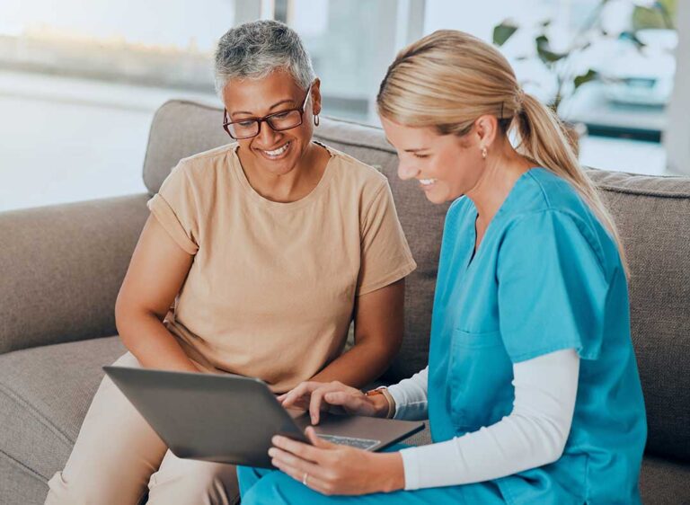 An older adult using a tablet device with a caregiver nearby in a cozy living room setting.