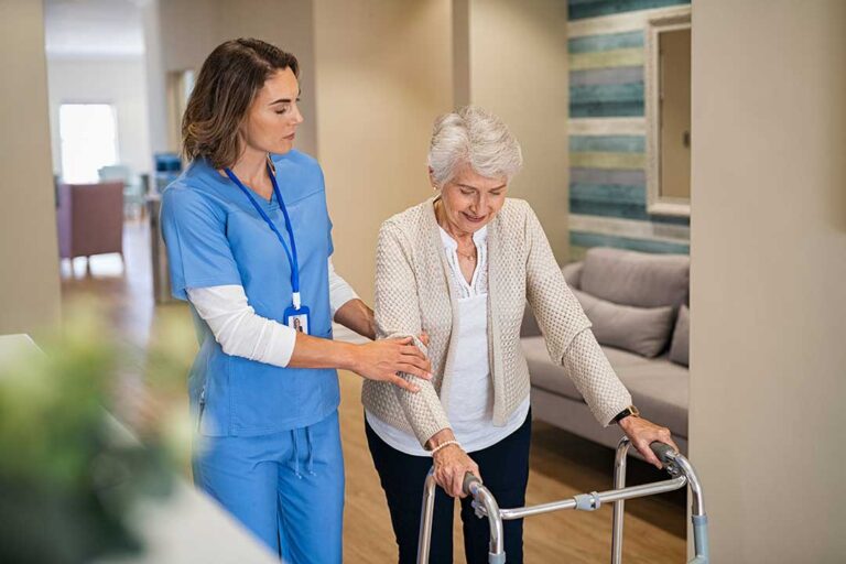 Older adult and family member organize labeled clothing, toiletries, photographs, and a walker in a bright assisted living room.